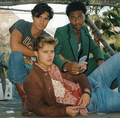 three young men sitting on the ground with one holding a cell phone and another looking at something in his hand