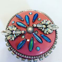 a pink and blue brooch with jewels on it's side, sitting on a white surface