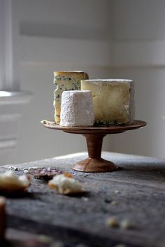 three different types of cheese on a wooden table with white walls in the background and food scattered around it