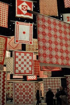 many red and white quilts are hanging from the ceiling in a room with people looking at them