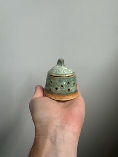 a hand holding a green ceramic jar with holes on it's lid, in front of a gray wall