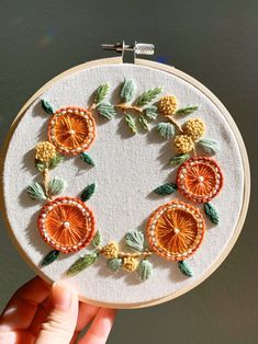 someone is holding up a small embroidery hoop with oranges and green leaves on it