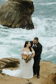 a bride and groom standing on the rocks by the ocean