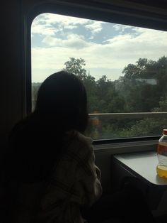 a person sitting on a train looking out the window at some trees and clouds in the distance