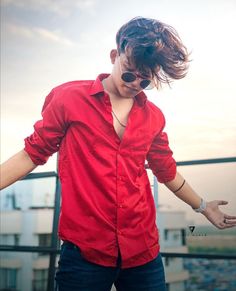 a man with his hair blowing in the wind on top of a roof, wearing sunglasses and a red shirt