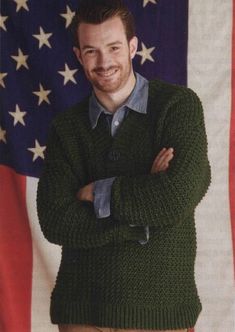 a man standing in front of an american flag wearing a green sweater and khaki pants