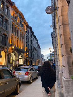 a woman is walking down the street in front of some cars and buildings at dusk