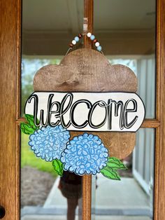 a welcome sign hanging on the front door of a house with blue hydrangeas