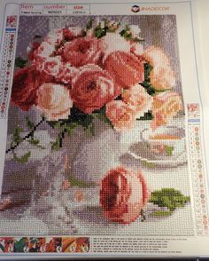 a cross stitch picture of pink roses in a white vase on a table with plates and cups