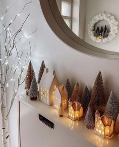 lighted christmas trees and small houses on a white shelf in front of a round mirror