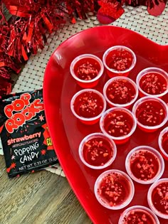 a heart shaped tray filled with cups and candy on top of a wooden table next to red tinsel