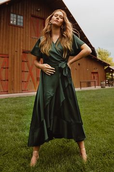 a woman standing in front of a barn with her hands on her hips wearing a green dress