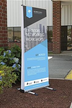 a sign advertising an exhibit in front of a brick building with blue flowers and bushes