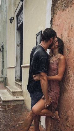 a man and woman leaning against a wall on a rainy day in the rain, kissing