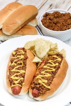 two chili dogs with mustard and potato chips on a white plate next to buns