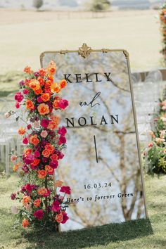 a sign with flowers on it in front of a tree and grass covered field behind it