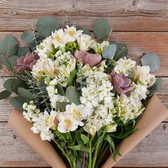 a bouquet of white flowers and greenery is wrapped in brown paper on a wooden surface