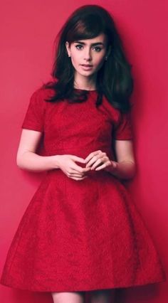 a woman in a red dress standing against a pink wall with her hands on her hips