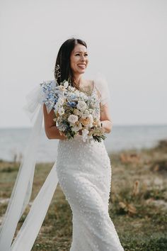 a woman in a wedding dress is holding a bouquet and walking on the grass near the water