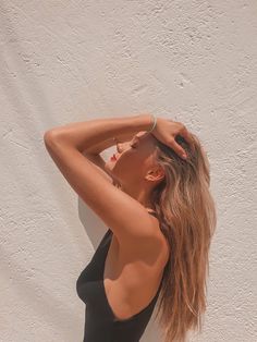 a woman in a black one piece swimsuit leaning against a wall with her arms behind her head