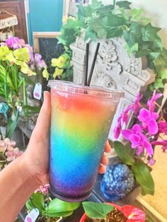 a hand holding a rainbow colored drink in front of flowers and other decorations on the table