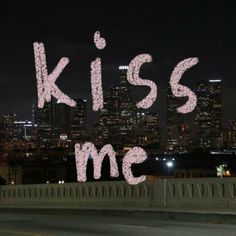 the words kiss me written in pink ink over a cityscape at night time