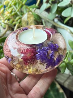 a hand holding a small glass candle holder with flowers in it and greenery behind it