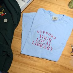 Show off your love for books and community with our "Support Your Local Library" embroidered shirt or crewneck. This cozy, customizable librarian sweatshirt is the perfect way to share your passion for reading and support the spaces that foster knowledge. Whether you're a proud book nerd or simply love curling up with a good read, this library lover tee makes a thoughtful gift for any literary enthusiast. Crafted for comfort and style, this bookworm t-shirt is perfect for anyone who values libra Library Shirt, Reading Day, Local Library, Gifts For Readers, Custom Embroidery, Librarian, Book Lovers Gifts, Embroidered Shirt, Book Nerd