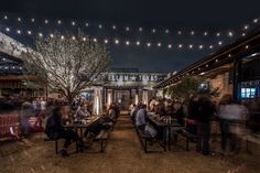 a group of people sitting at tables in the middle of an outdoor area with string lights