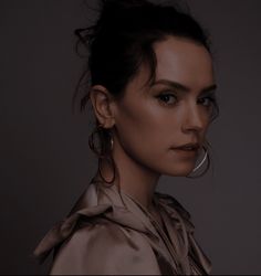 a woman with large hoop earrings on her ear and wearing a silver shirt, standing in front of a dark background