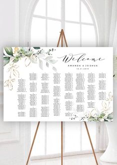 a white and gold wedding seating chart with greenery on the side, in front of a window