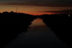 the sun is setting over a canal with trees on both sides and hills in the distance