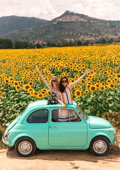 two women standing on top of a car in front of a field of sunflowers