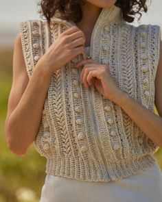 a woman wearing a white knitted vest and holding her hand on her chest with both hands