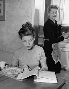 two women are sitting at a kitchen table and one is writing in a book while the other woman looks on