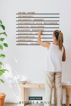 a woman standing in front of a white wall with writing on it and a potted plant behind her