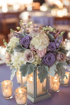 purple and white flowers are in a silver vase on a blue tablecloth with candles