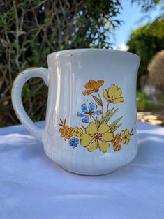 a white coffee cup with yellow and blue flowers on it sitting on a table outside