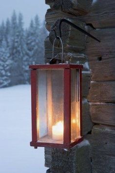 a lantern hanging from the side of a log cabin in the snow with two lit candles inside