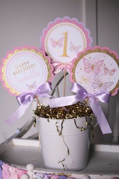 a white bucket filled with pink and gold birthday cupcake toppers next to a purple ribbon