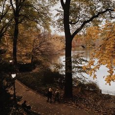 two people walking down a path next to a lake in the fall with leaves on the ground