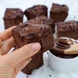 a hand holding a piece of chocolate brownie in front of some cupcakes