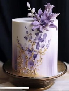 a white and purple cake with flowers on the top is sitting on a gold plate