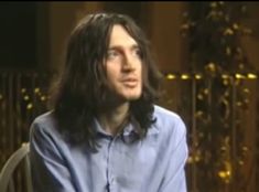 a man with long hair sitting in a chair and looking off to the side while wearing a blue shirt