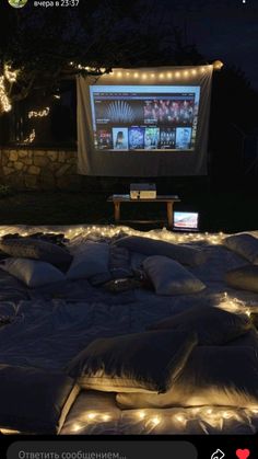 an outdoor movie screen is lit up with fairy lights and pillows on the ground in front of it