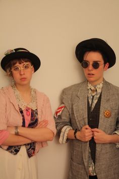 two people wearing hats and glasses standing next to each other in front of a white wall