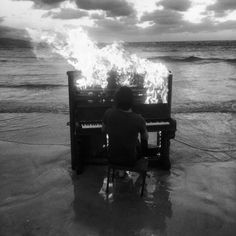 a man sitting in front of a piano on top of a beach next to the ocean