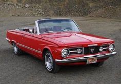 an old red convertible car parked in a parking lot next to a stone wall and dirt area