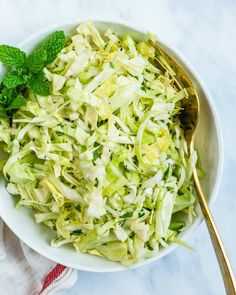 a white bowl filled with coleslaw next to a gold spoon and green leafy garnish