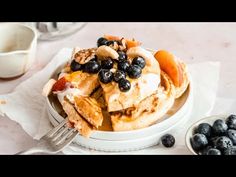 a white plate topped with pancakes covered in blueberries and peaches next to a bowl of fruit